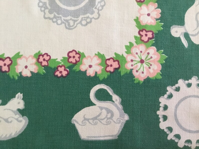 May include: A green fabric with a white and pink floral pattern. The fabric features a white swan, a white rabbit, and a white plate with a lace border.
