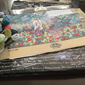 May include: A needlepoint kit featuring a woman in a straw hat walking through a field of red poppies. The kit includes DMC embroidery floss in various colors and a beige canvas with a 40 x 30 count. The packaging includes the text "On trouve tout Paris a la Samaritaine."