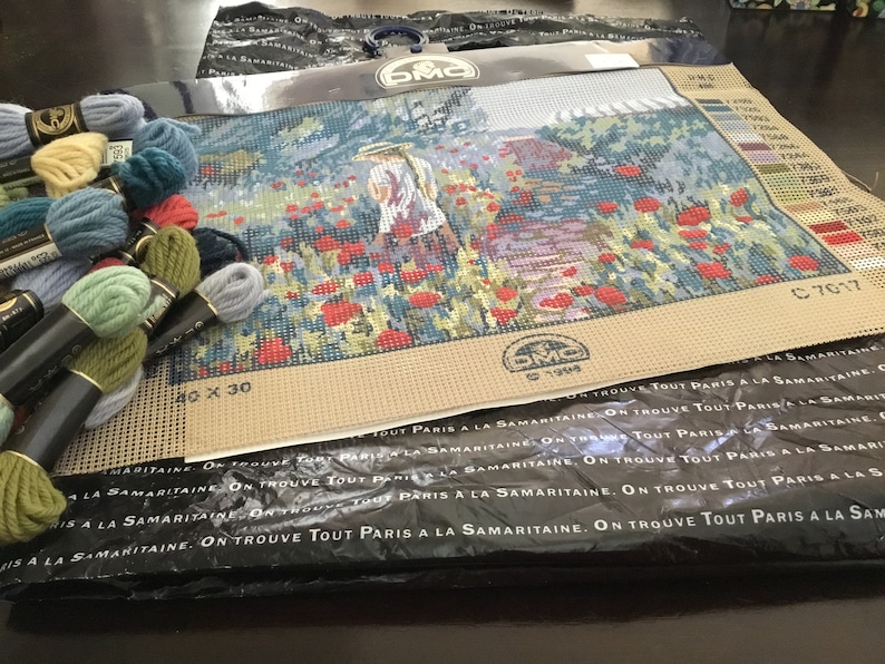 May include: A needlepoint kit featuring a woman in a straw hat walking through a field of red poppies. The kit includes DMC embroidery floss in various colors and a beige canvas with a 40 x 30 count. The packaging includes the text "On trouve tout Paris a la Samaritaine."