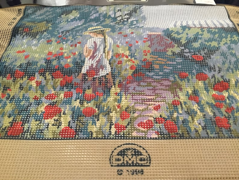May include: A needlepoint canvas with a girl in a white dress and a straw hat standing in a field of red poppies. The canvas is beige with a brown border and a logo in the bottom center.