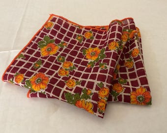 May include: Folded burgundy handkerchief with a white grid pattern and orange floral accents. The handkerchief has an orange border and is made of a lightweight fabric. The floral design includes orange and yellow flowers with green leaves.