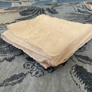 May include: A white cotton handkerchief with a delicate floral embroidery design. The handkerchief is folded and placed on a blue and white patterned fabric.
