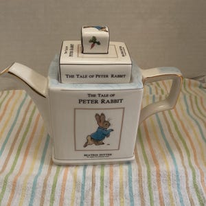 May include: A square white ceramic teapot featuring The Tale of Peter Rabbit illustrations. The lid has a small knob with a carrot graphic. The body displays the title, a rabbit in a blue jacket, and the Beatrix Potter signature in black text.