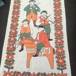 May include: A white tea towel with a red, orange, and green folk art design featuring four figures playing musical instruments and a person riding a wooden horse. The border is a repeating pattern of red flowers with green leaves. The text "ULLAS." is in the bottom right corner.