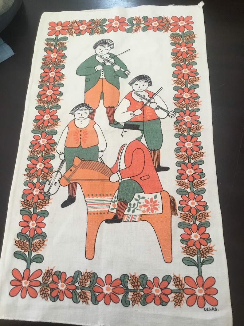 May include: A white tea towel with a red, orange, and green folk art design featuring four figures playing musical instruments and a person riding a wooden horse. The border is a repeating pattern of red flowers with green leaves. The text "ULLAS." is in the bottom right corner.
