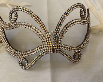 May include: A decorative mask with a butterfly design. The mask is covered in small, iridescent rhinestones, with a larger rhinestone at the center. The frame is a dark brown color, and the mask has a delicate, elegant appearance.