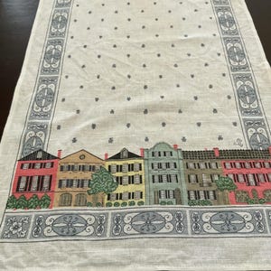 May include: Cream-colored table runner with a repeating pattern of small dark blue floral shapes. The bottom edge features a row of colorful buildings with red, yellow, green, and brown facades. The border has a decorative blue and white design.