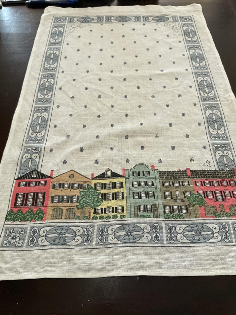 May include: Cream-colored table runner with a repeating pattern of small dark blue floral shapes. The bottom edge features a row of colorful buildings with red, yellow, green, and brown facades. The border has a decorative blue and white design.