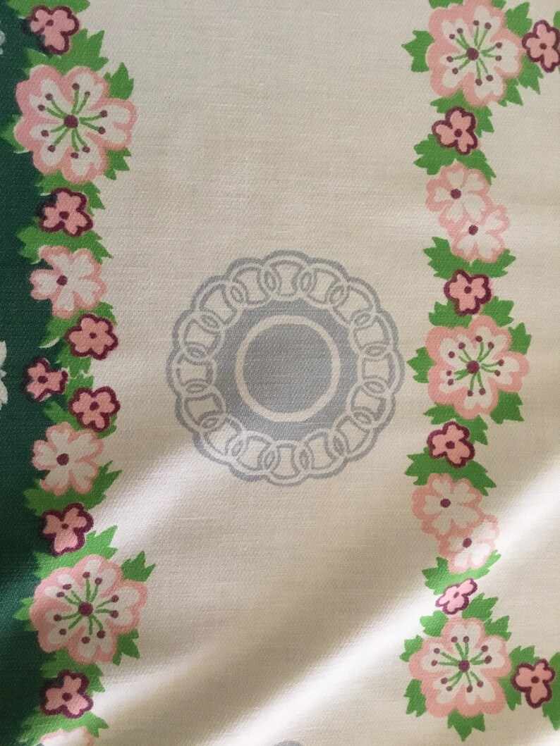 May include: A white tablecloth with a floral pattern in shades of pink, green, and grey. The tablecloth features a circular design in the centre with a grey background and a white border.