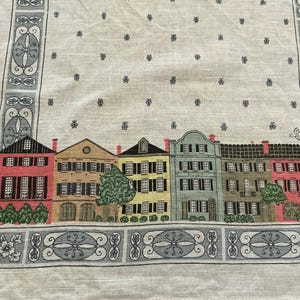 May include: A white linen table runner featuring a row of colorful houses with black shutters and red brick chimneys. The runner has a decorative border with floral and geometric patterns, and small blue floral accents.