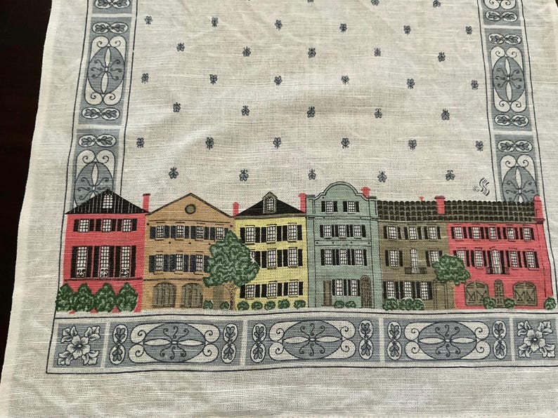May include: A white linen table runner featuring a row of colorful houses with black shutters and red brick chimneys. The runner has a decorative border with floral and geometric patterns, and small blue floral accents.