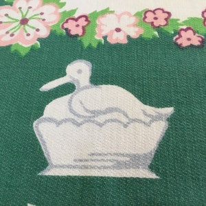 May include: A white duck with a grey beak sitting in a white nest on a green background with pink flowers.