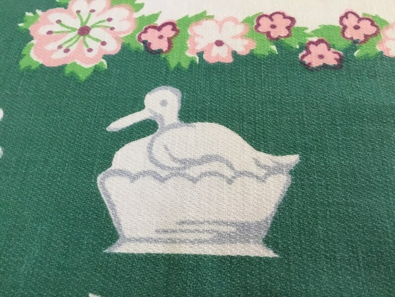 May include: A white duck with a grey beak sitting in a white nest on a green background with pink flowers.