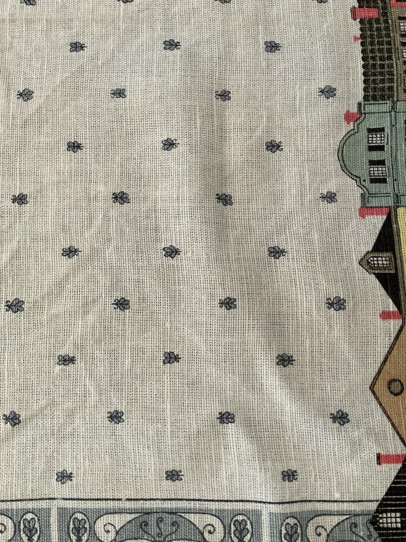 May include: Cream-colored fabric with a repeating pattern of small, dark blue floral motifs. The right edge features a partial illustration of buildings in various colors, including green, brown, and black. A decorative border runs along the bottom.