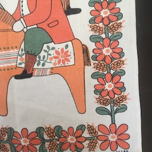 May include: A vintage white linen tea towel with a colorful folk art design of a person riding a wooden horse. The design features orange, red, green, and black colors. The towel has a border of orange flowers and green leaves. The text "ULLAS." is printed in the bottom right corner.