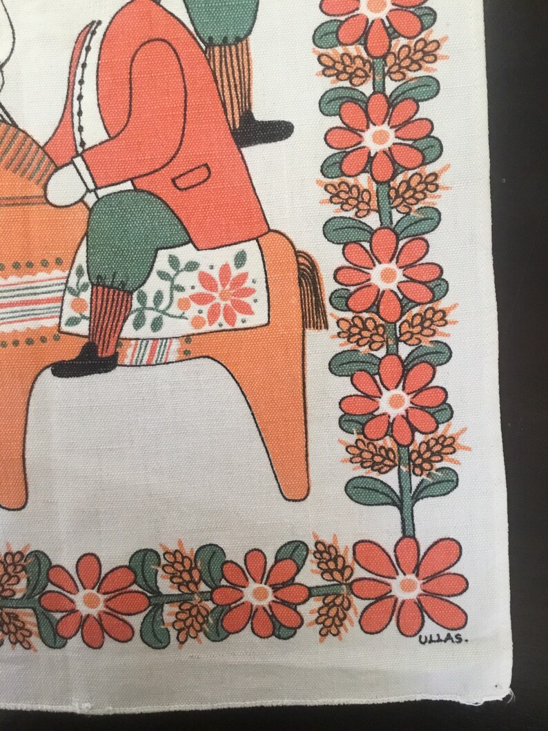 May include: A vintage white linen tea towel with a colorful folk art design of a person riding a wooden horse. The design features orange, red, green, and black colors. The towel has a border of orange flowers and green leaves. The text "ULLAS." is printed in the bottom right corner.