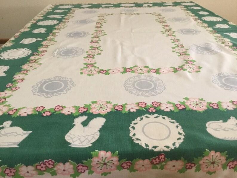 May include: A white tablecloth with a green border featuring a repeating pattern of pink flowers, white chickens in nests, and white plates. The tablecloth has a white centre with a repeating pattern of grey plates.