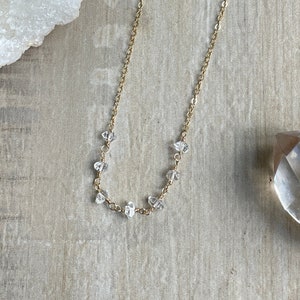 Pode incluir: Um colar de corrente de ouro com detalhes de cristal transparente. O colar está sobre uma superfície de madeira marrom claro.