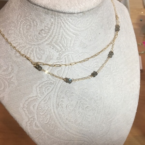 May include: A gold-colored, layered necklace with small, gray-green gemstones. The necklace is displayed on a white, fabric-covered bust. The necklace has two strands, one with a paperclip chain design and the other with a delicate chain.