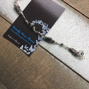 Może przedstawiać: Ręcznie robiona bransoletka z ciemnymi, prostokątnymi koralikami owiniętymi srebrnym drutem. Obok czarna wizytówka z motywem motyla i napisem "Butterfly Kisses Hawaii". Na drewnianej powierzchni.