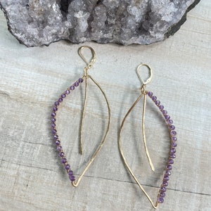 Könnte beinhalten: Ein Paar goldfarbene, blattförmige Ohrringe. Die Ohrringe haben ein filigranes Design mit einer Reihe kleiner, lila Perlen am äußeren Rand. Die Ohrringe haben einen Hakenverschluss und sind auf Holz präsentiert.