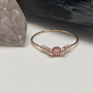 May include: A delicate gold ring with a single pink gemstone wrapped in wire.