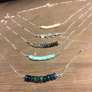 Pode incluir: Cinco colares de prata com contas de pedras preciosas de diferentes cores. Os colares estão sobrepostos numa superfície de madeira. O colar superior tem contas cor-de-rosa, o segundo tem contas transparentes e verdes, o terceiro tem contas azuis e verdes, o quarto tem contas turquesa e o colar inferior tem contas azul escuro e verdes.
