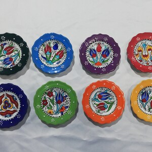 May include: Eight small decorative ceramic plates with scalloped edges. Each plate features a different color background and a hand-painted floral design with blue, red, yellow, and green colors.