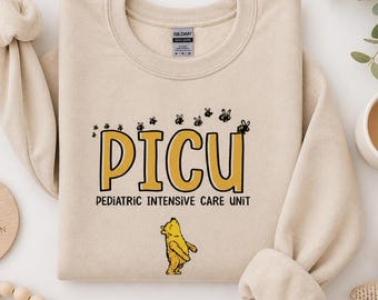 May include: A beige sweatshirt with the text "PICU Pediatric Intensive Care Unit" in yellow and black. The design includes cartoon bees and a Winnie the Pooh character. The sweatshirt is folded with the sleeves cuffed.