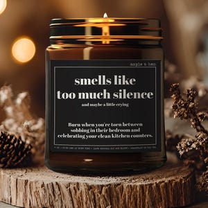 May include: A lit candle in a brown glass jar with a black label that reads "smells like too much silence." The candle sits on a wooden slice, surrounded by dried plants and bokeh lights. The jar has a gold-colored lid.