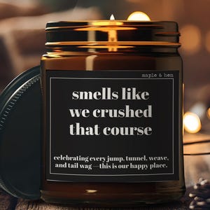 May include: A lit brown glass jar candle with a black lid, featuring a black label with white text that reads "smells like we crushed that course." The candle is on a wooden surface with a blurred background.
