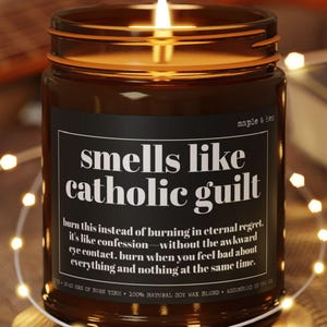 May include: A brown glass candle with a black label that reads 'smells like catholic guilt'. The label also includes the text 'burn this instead of burning in eternal regret. it's like confession without the awkward eye contact. burn when you feel bad about everything and nothing at the same time.' The candle is lit and sitting on a wooden coaster.