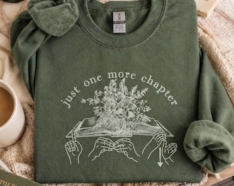 May include: Olive green sweatshirt with the text "just one more chapter" and a floral book illustration. The image includes books, a candle, a mug, and reading glasses, suggesting a cozy reading theme.