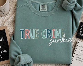 May include: A sage green sweatshirt with the text "TRUE CRIME junkie" in colorful letters. The image includes a mug of coffee, a small chalkboard with the words "STAY CURIOUS STAY CAREFUL", and a notepad with a podcast list.