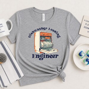 May include: A gray t-shirt with a graphic of a dishwasher and the words "Dishwasher Loading Engineer." A white mug with the text "Everything Has A Place, Even This Mug" is next to a striped towel and a dish brush.