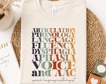 May include: A cream-coloured t-shirt featuring a colourful text design listing speech pathology terms. The words include Articulation, Phonology, Language, Fluency, Dysphagia, Aphasia, Voice, and AAC. The text "speech language pathologist" is at the bottom.