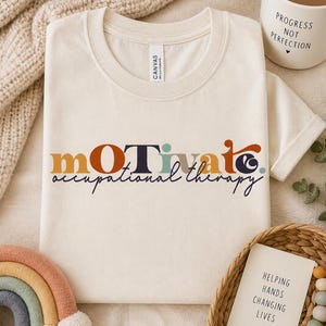 May include: Cream-colored t-shirt with the words "MOTIVATE occupational therapy" in colorful lettering. A white mug with the words "PROGRESS NOT PERFECTION" is visible. A rainbow-shaped toy and a woven basket with a card are also in the image.