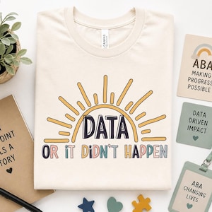 May include: Cream-colored t-shirt with a sun graphic and the text "DATA OR IT DIDN'T HAPPEN". The image includes a notebook, cards with text, and small wooden shapes. The shirt is a casual wear item.