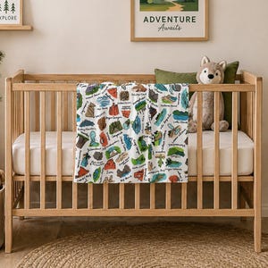 May include: A wooden crib with a white mattress and a colorful blanket featuring various national park illustrations. A framed print that says "Adventure Awaits" hangs above the crib. A stuffed wolf and green pillow are inside the crib. A small wooden sign that says "Let's Explore" is on a shelf.