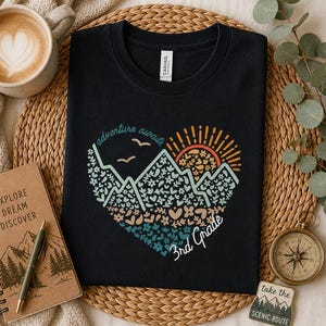 May include: Black t-shirt with a heart-shaped mountain and sun design. The words "adventure awaits" and "3rd Grade" are printed on the shirt. A coffee cup, notebook, pen, compass, and coaster are arranged around the shirt.