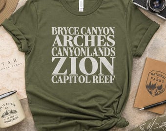 Könnte beinhalten: Ein olivgrünes T-Shirt mit weißem Text, der die Namen von fünf Nationalparks auflistet: Bryce Canyon, Arches, Canyonlands, Zion und Capitol Reef. Das T-Shirt hat einen Rundhalsausschnitt und kurze Ärmel, wobei der Text mittig auf der Brust platziert ist.