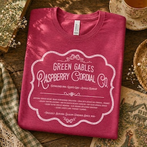May include: A crimson t-shirt with a crew neck, featuring a white graphic that reads "Green Gables Raspberry Cordial Co." The design includes decorative flourishes and text detailing additional services. The shirt is folded on a wooden surface.
