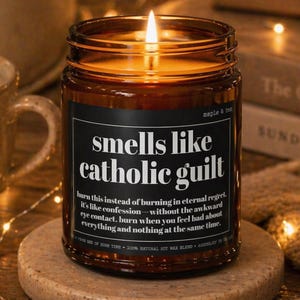 May include: A lit brown glass candle jar with the text "smells like catholic guilt" on a black label. The candle sits on a round stone coaster, with a cup of coffee and a decorative bowl in the background. The candle is surrounded by fairy lights.