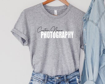 May include: A grey t-shirt with the text "Custom Name PHOTOGRAPHY" printed on it. The t-shirt is hanging on a wooden hanger and is paired with a light blue denim shirt.