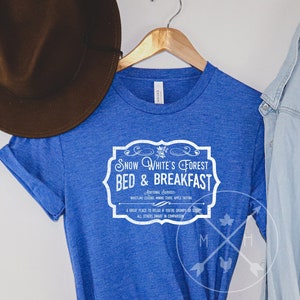 May include: A royal blue t-shirt with a white graphic design that reads "Snow White's Forest Bed & Breakfast". The shirt is on a hanger with a brown hat and a light blue denim jacket.