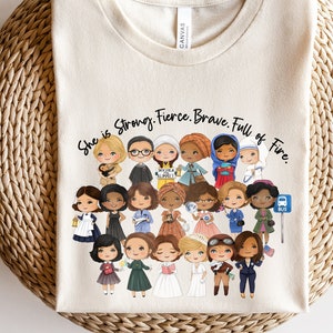 Pu&ograve; includere: T-shirt color crema con un gruppo di donne illustrate di diverse etnie e professioni. Il testo "She is Strong. Fierce. Brave. Full of Fire" &egrave; stampato sopra le illustrazioni. La maglietta &egrave; realizzata in un materiale morbido.