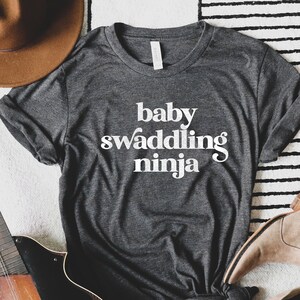 Puede incluir: Camiseta gris oscuro con la frase "baby swaddling ninja" en blanco. La imagen incluye un sombrero marr&oacute;n, una mandolina, botas color beige y un peque&ntilde;o cactus en una maceta rosa, sobre una alfombra.