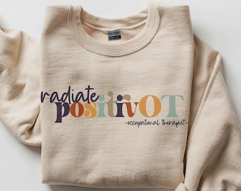 Op de afbeelding: Een beige sweatshirt met de tekst "radiate positivity OT" in een kleurrijk retro-lettertype. Onder de tekst staat de zin "occupational therapist". De sweatshirt is opgevouwen met de mouwen aan de zijkant.