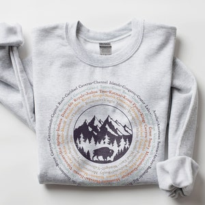 May include: A light grey crewneck sweatshirt featuring a circular graphic on the chest. The design includes a dark purple silhouette of mountains, pine trees, and a bison, surrounded by a ring of text listing various national park names.