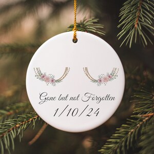 May include: White ceramic ornament with a gold string, featuring a floral design with pink flowers and green leaves. The ornament has the text "Gone but not Forgotten 1/10/24" written on it.